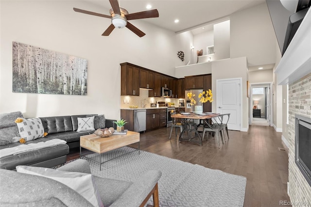 living room featuring ceiling fan, sink, a fireplace, a towering ceiling, and dark hardwood / wood-style flooring