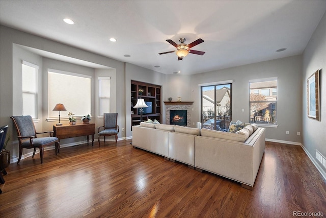 living room featuring a stone fireplace, built in features, dark hardwood / wood-style floors, and ceiling fan