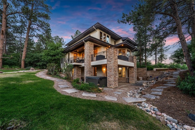 back house at dusk featuring a hot tub, a balcony, and a yard