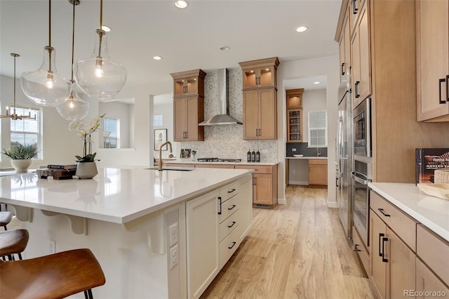 kitchen with wall chimney range hood, an island with sink, sink, a kitchen bar, and light hardwood / wood-style floors