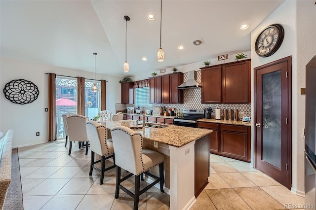 kitchen featuring stainless steel range with electric stovetop, a center island with sink, wall chimney exhaust hood, decorative light fixtures, and decorative backsplash