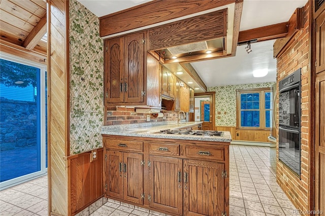 kitchen with kitchen peninsula, beam ceiling, stainless steel gas cooktop, double oven, and light tile patterned floors