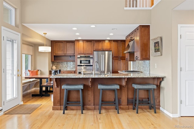 kitchen featuring light stone counters, under cabinet range hood, a peninsula, appliances with stainless steel finishes, and brown cabinets