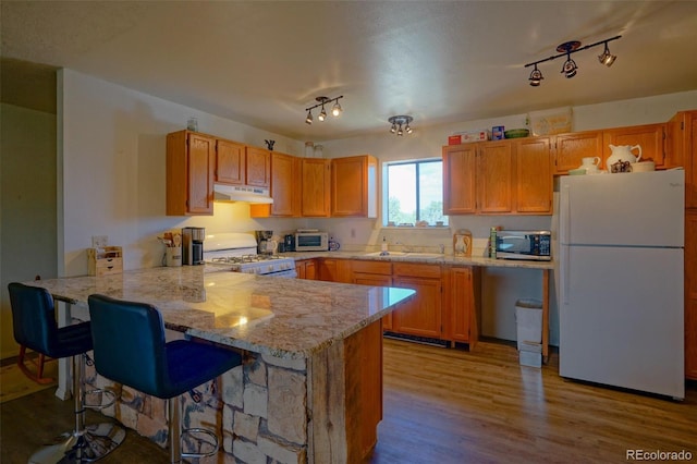 kitchen featuring wood finished floors, a kitchen bar, a peninsula, under cabinet range hood, and white appliances