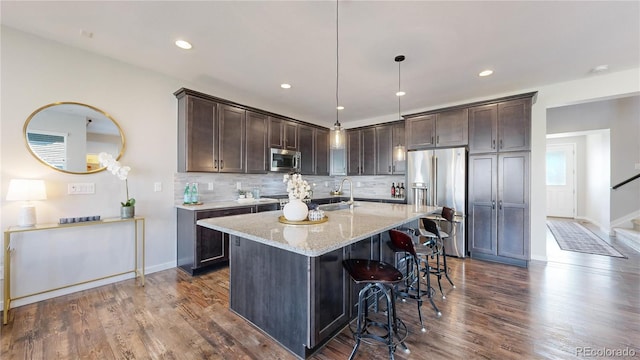 kitchen featuring stainless steel appliances, a kitchen breakfast bar, light stone countertops, dark wood-type flooring, and a center island with sink