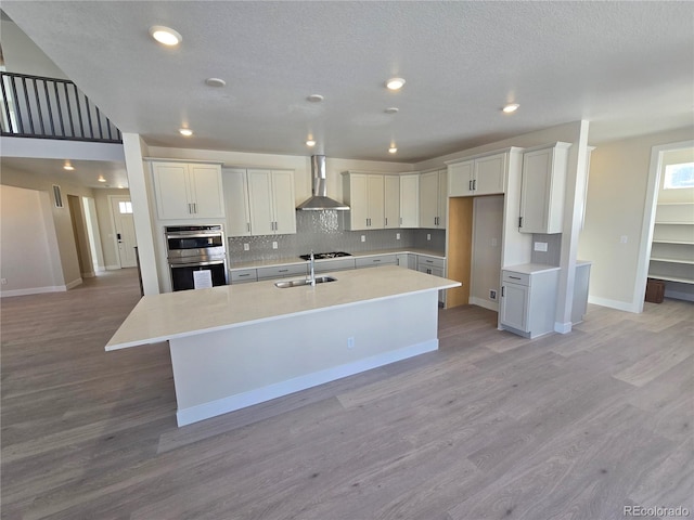 kitchen with wall chimney range hood, light hardwood / wood-style floors, a kitchen island with sink, white cabinets, and appliances with stainless steel finishes