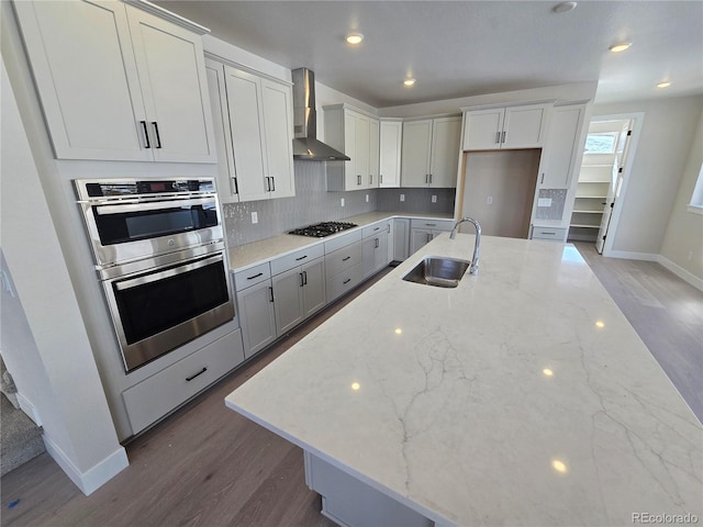 kitchen with sink, wall chimney exhaust hood, light stone counters, wood-type flooring, and appliances with stainless steel finishes