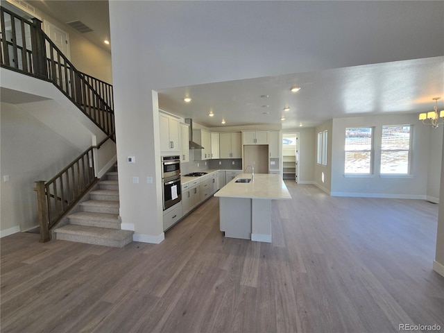 kitchen featuring a kitchen island with sink, hardwood / wood-style flooring, wall chimney exhaust hood, appliances with stainless steel finishes, and white cabinetry