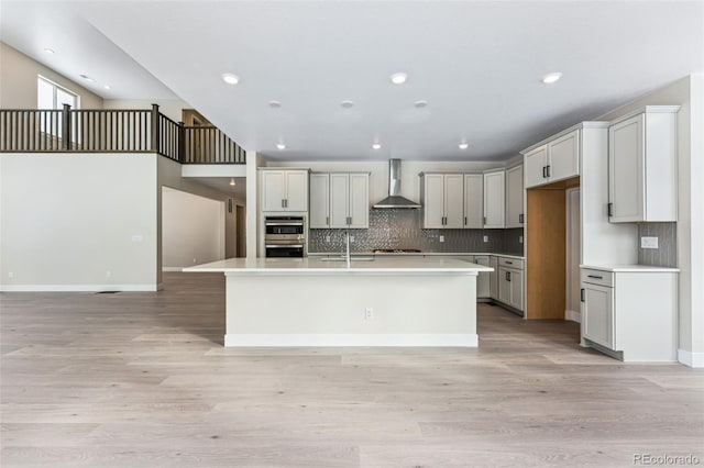 kitchen with a center island with sink, light hardwood / wood-style flooring, wall chimney exhaust hood, decorative backsplash, and gas stovetop