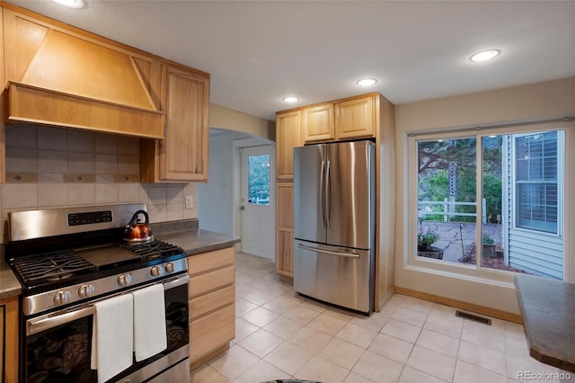 kitchen with light tile patterned flooring, light brown cabinetry, custom exhaust hood, stainless steel appliances, and decorative backsplash