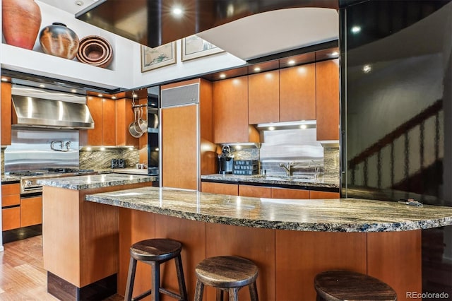 kitchen featuring brown cabinets, dark stone counters, and wall chimney range hood