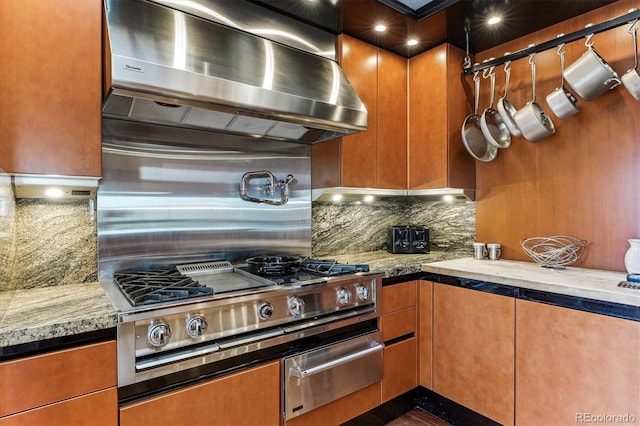 kitchen featuring light stone countertops, decorative backsplash, brown cabinetry, wall chimney exhaust hood, and a warming drawer