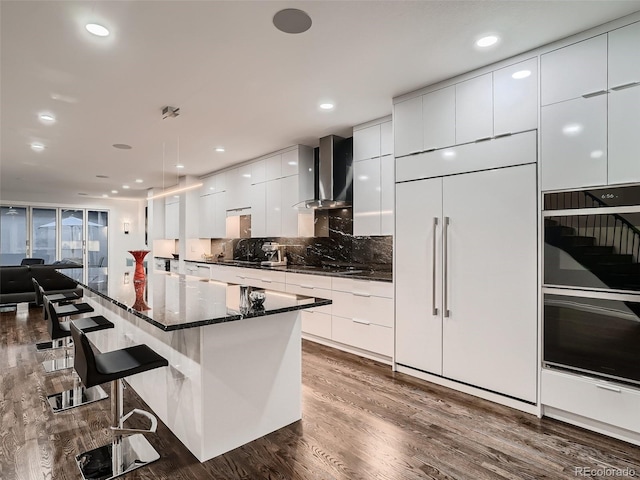 kitchen with dobule oven black, modern cabinets, a breakfast bar, wall chimney exhaust hood, and dark wood-type flooring