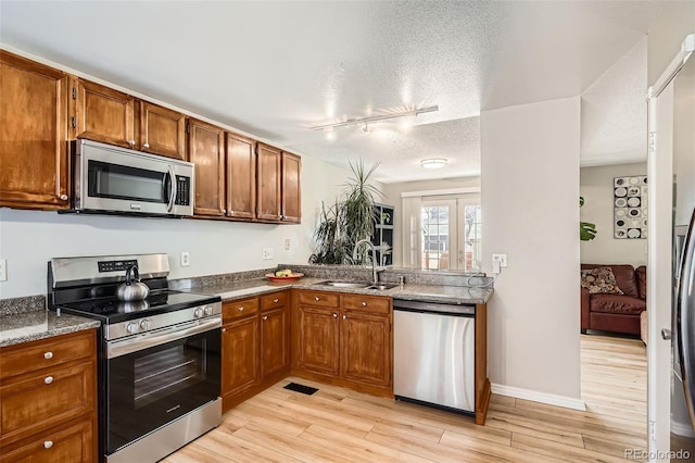 kitchen with brown cabinetry, light wood-style flooring, a sink, appliances with stainless steel finishes, and a textured ceiling