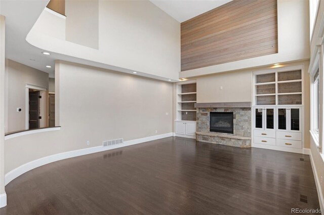 unfurnished living room with built in shelves, a towering ceiling, dark hardwood / wood-style flooring, and a stone fireplace