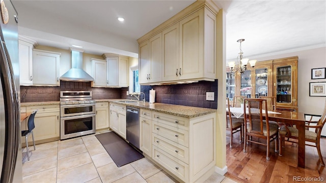 kitchen with wall chimney exhaust hood, tasteful backsplash, appliances with stainless steel finishes, cream cabinets, and light hardwood / wood-style floors