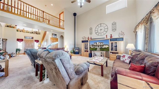 carpeted living room with ceiling fan, a towering ceiling, a wood stove, and plenty of natural light