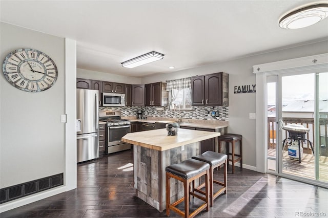 kitchen featuring a kitchen bar, stainless steel appliances, butcher block counters, decorative backsplash, and dark brown cabinets