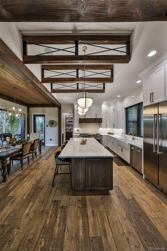 kitchen featuring stainless steel appliances, a kitchen island, dark wood-type flooring, light stone countertops, and beam ceiling