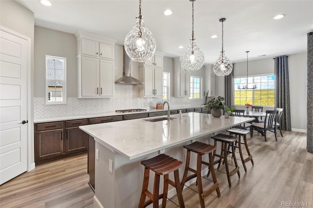 7000 S White Crow Way, Aurora, CO 80016 - kitchen featuring decorative backsplash, an island with sink, recessed lighting, light wood-type flooring, and wall chimney exhaust hood
