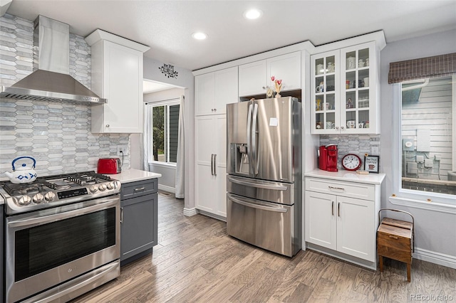 kitchen featuring tasteful backsplash, stainless steel appliances, wall chimney range hood, hardwood / wood-style flooring, and white cabinets