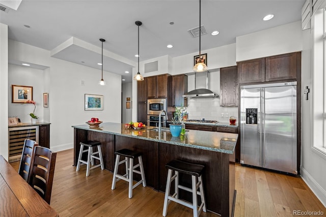 kitchen featuring built in appliances, wall chimney exhaust hood, light wood-type flooring, an island with sink, and a breakfast bar area