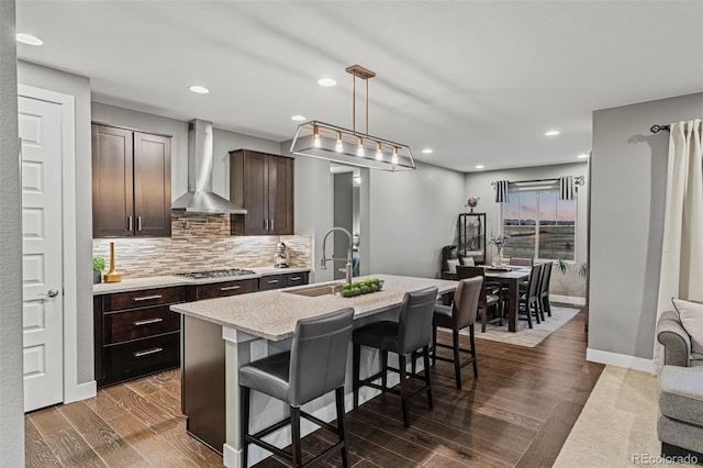 kitchen featuring a center island with sink, dark brown cabinets, a kitchen breakfast bar, wall chimney range hood, and sink