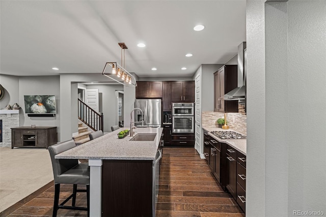 kitchen with a center island with sink, stainless steel appliances, a breakfast bar, hanging light fixtures, and wall chimney range hood