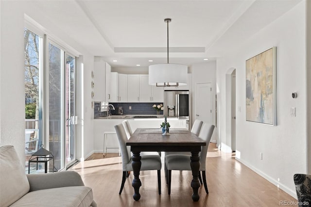 dining room with arched walkways, baseboards, light wood-style flooring, a tray ceiling, and recessed lighting