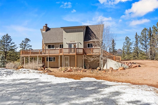 snow covered house featuring a wooden deck, stucco siding, roof with shingles, and a chimney