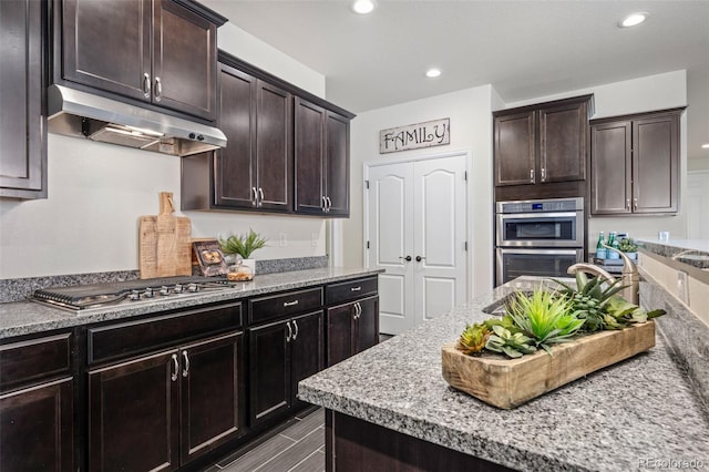 kitchen with appliances with stainless steel finishes, light stone counters, dark brown cabinets, under cabinet range hood, and recessed lighting