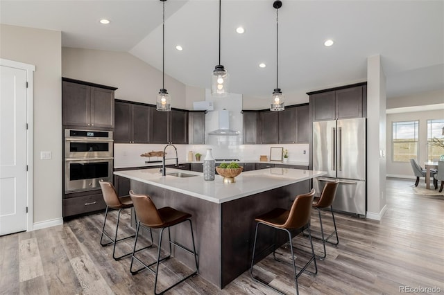 kitchen with sink, wall chimney range hood, a kitchen island with sink, stainless steel appliances, and decorative light fixtures