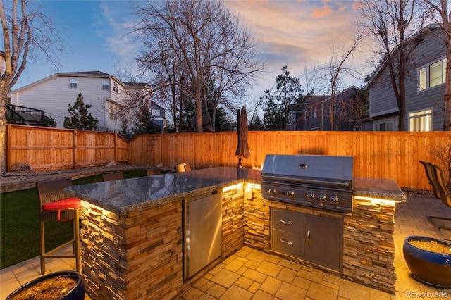 patio terrace at dusk with exterior kitchen, a fenced backyard, and a grill