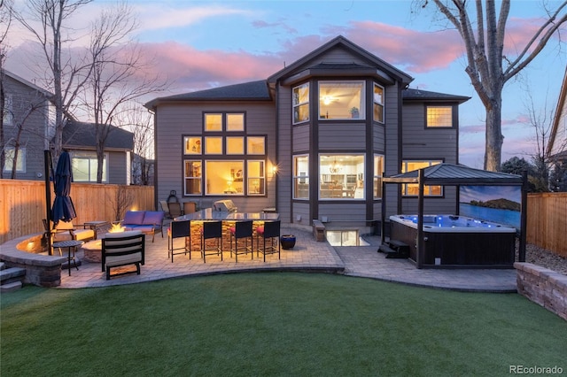 back of property at dusk featuring a fenced backyard, a fire pit, outdoor dry bar, a patio area, and a hot tub