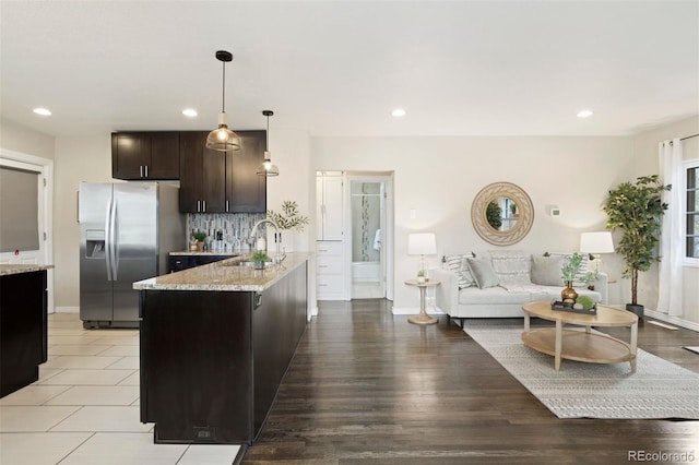 kitchen featuring light stone countertops, stainless steel fridge, dark brown cabinetry, pendant lighting, and a center island with sink