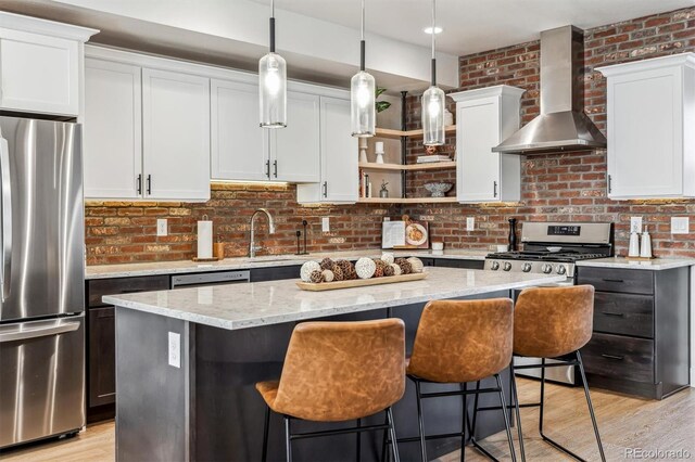 kitchen with brick wall, appliances with stainless steel finishes, a center island, wall chimney range hood, and open shelves