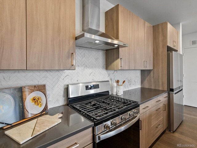 kitchen featuring light brown cabinetry, dark countertops, stainless steel appliances, and wall chimney exhaust hood