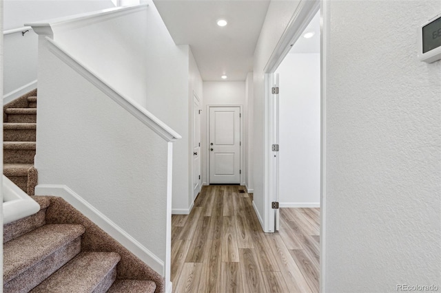 hall featuring a textured wall, recessed lighting, baseboards, stairs, and light wood-style floors