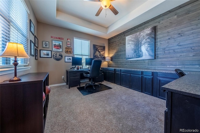 carpeted office space featuring a tray ceiling, wood walls, and ceiling fan