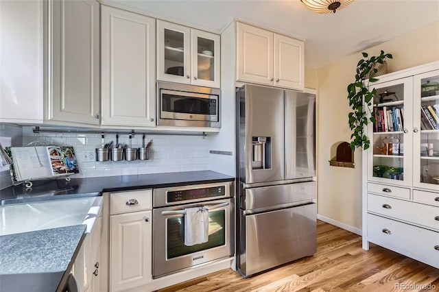 kitchen featuring light wood-type flooring, white cabinets, glass insert cabinets, appliances with stainless steel finishes, and tasteful backsplash