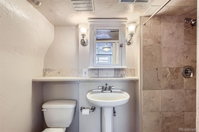 bathroom featuring visible vents, toilet, a sink, tiled shower, and a textured wall