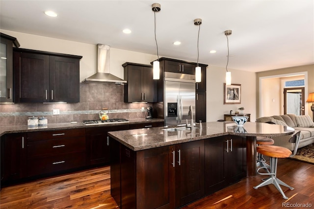 kitchen featuring dark wood-type flooring, appliances with stainless steel finishes, a kitchen bar, wall chimney range hood, and backsplash