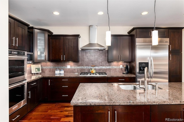 kitchen featuring dark brown cabinets, wall chimney range hood, decorative backsplash, stainless steel appliances, and a sink