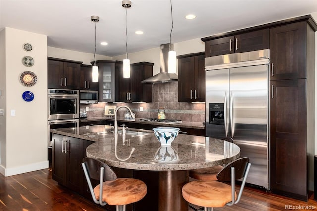 kitchen with a sink, dark brown cabinets, appliances with stainless steel finishes, wall chimney range hood, and tasteful backsplash