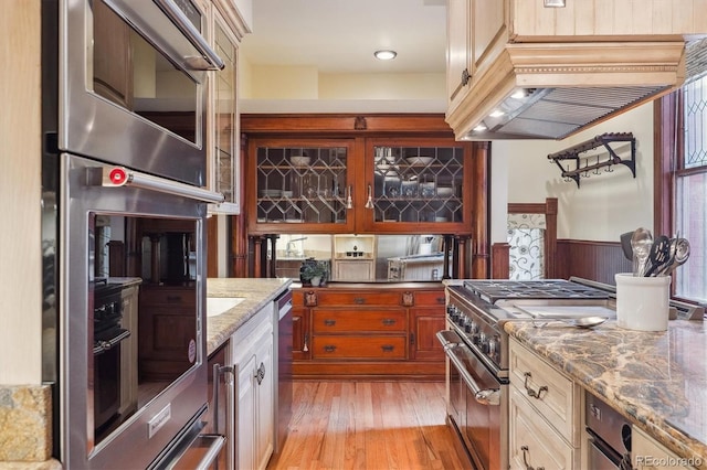 kitchen featuring appliances with stainless steel finishes, wainscoting, light stone counters, light wood-style flooring, and glass insert cabinets