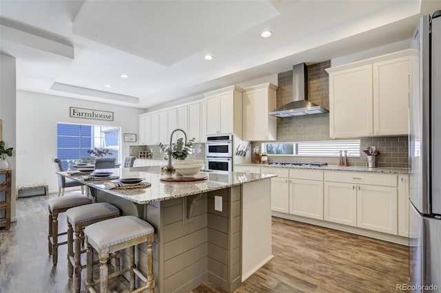 kitchen with wall chimney exhaust hood, wood finished floors, appliances with stainless steel finishes, tasteful backsplash, and recessed lighting