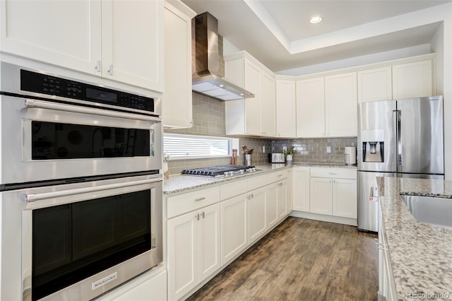 kitchen with wall chimney exhaust hood, stainless steel appliances, white cabinets, wood finished floors, and tasteful backsplash