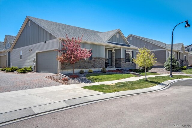 craftsman-style house with an attached garage, decorative driveway, a front lawn, stone siding, and roof with shingles