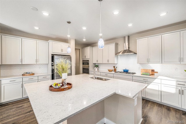 kitchen featuring a center island with sink, dark wood finished floors, a sink, appliances with stainless steel finishes, and wall chimney range hood