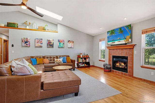 living room with ceiling fan, light hardwood / wood-style floors, vaulted ceiling with skylight, and a tile fireplace